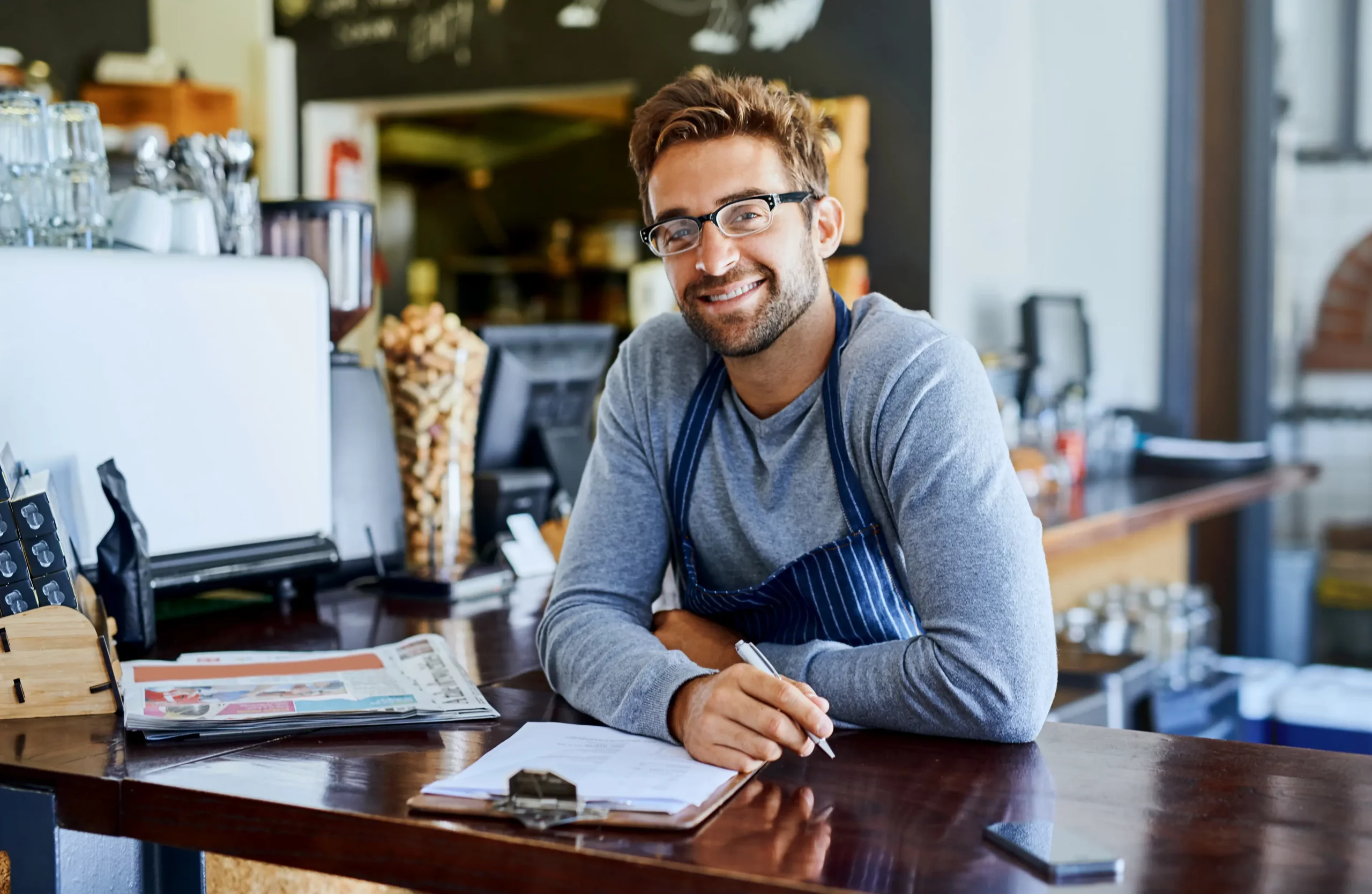 A smiling young business owner of a cafe, wearing glasses and a striped apron over a grey sweater, sits at a wooden counter with a clipboard and pen in hand, surrounded by coffee equipment and fresh pastries—exploring the key question: Can a Sole Trader Have Employees?