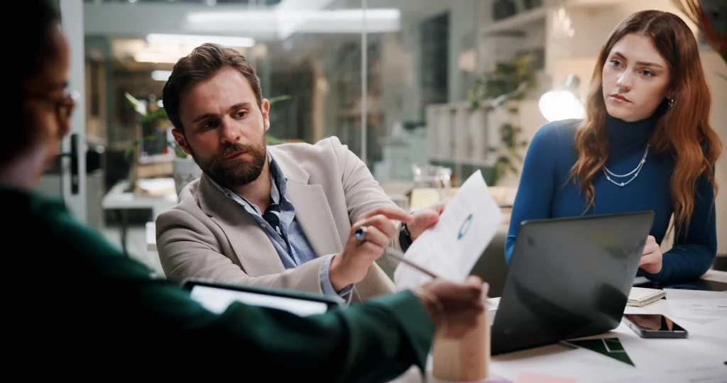 Three professionals in a modern office conducting a disciplinary hearing, with one manager pointing to documents while discussing with colleagues around a table—essential guide on how to conduct a disciplinary hearing fairly and compliantly.