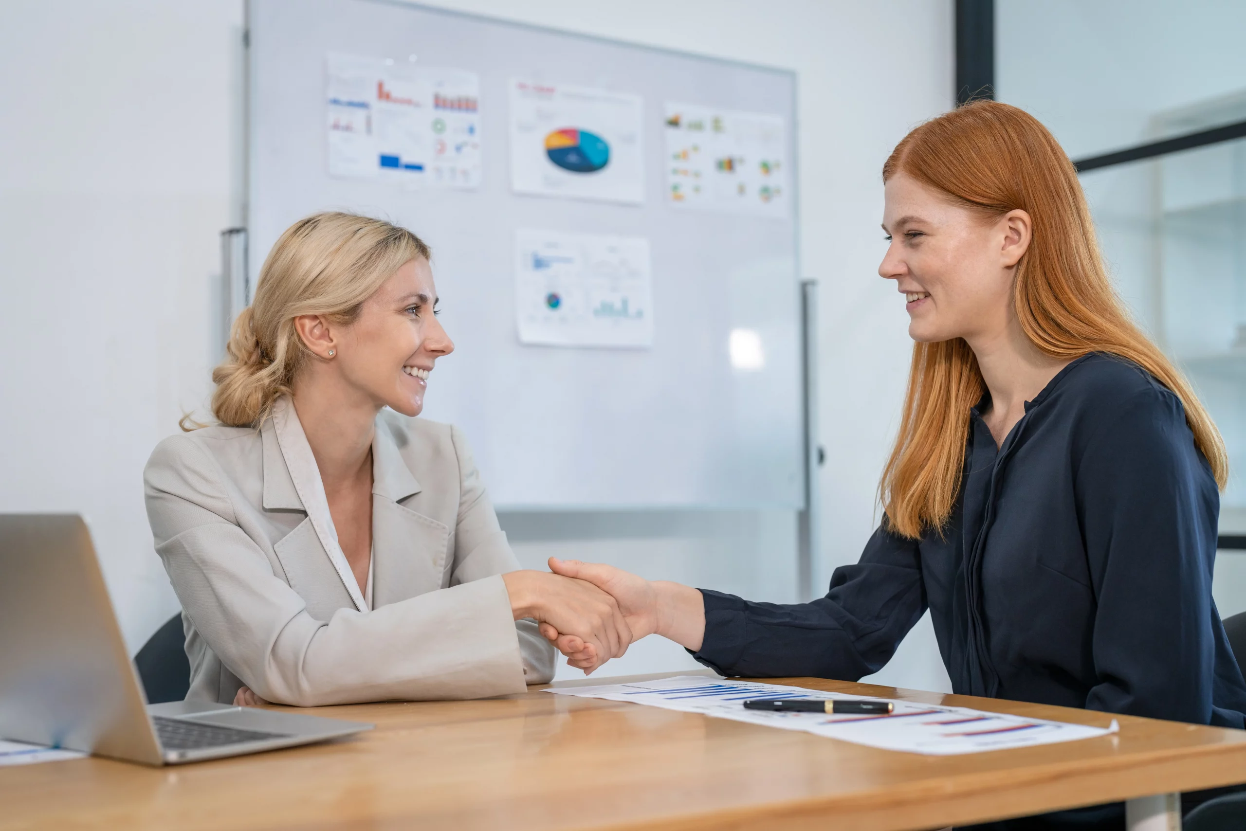 Two professional women shaking hands across a conference table in a modern office, demonstrating how to conduct a welfare meeting with empathy and collaboration, with charts on a whiteboard and documents nearby.