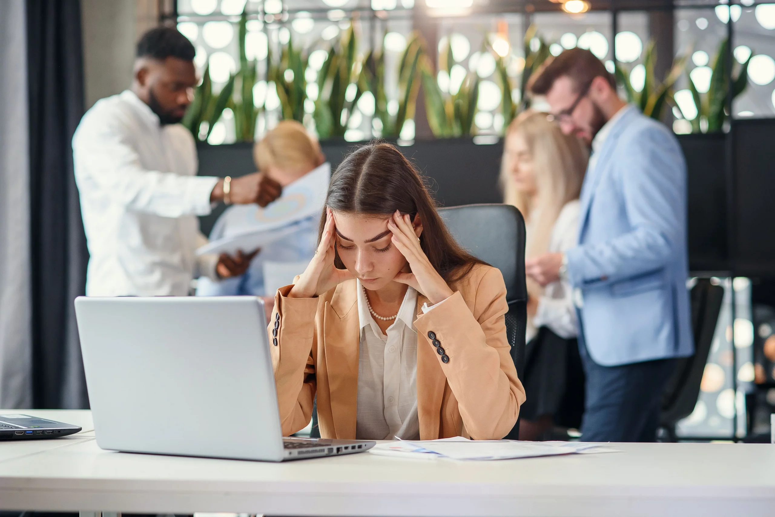 A stressed young woman in a beige blazer sits at a desk in a modern office, clutching her head with both hands in apparent anxiety, while blurred colleagues collaborate nearby amid plants and windows—highlighting the real-world challenges of managing staff with anxiety issues.