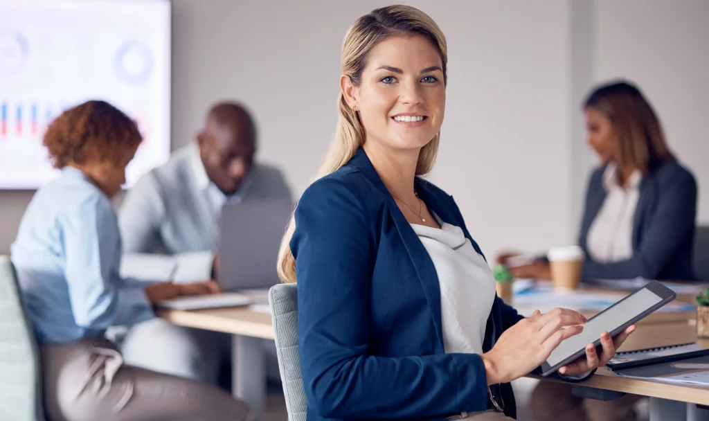 Expert executive recruitment agency specialising in exec recruitment agency and retained recruitment agency solutions: Confident blonde professional in navy blazer holding tablet, smiling amid diverse colleagues at modern office desks with laptops, coffee cups, charts on screen, and plants, fostering strategic senior talent acquisition.