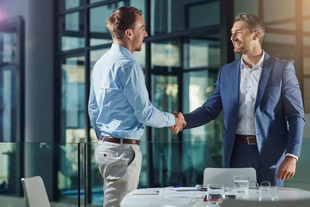 Professional recruitment consulting from a trusted recruitment agency among leading recruitment agencies: Two smiling businessmen in blue shirts and suits firmly shaking hands at modern glass-walled office desk with documents, pens, and water glasses, embodying successful talent acquisition and partnership agreements.
