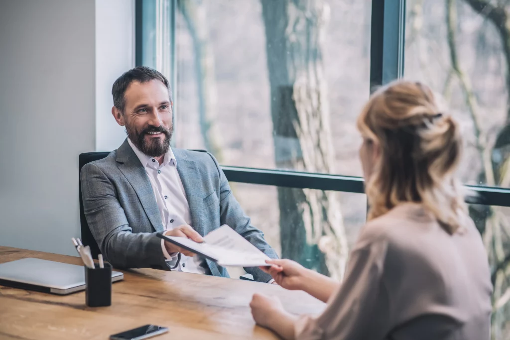 Professional recruitment consultant from trusted recruitment agency among top recruitment agencies: Smiling male expert in gray suit jacket handing documents to female job candidate in beige blouse at wooden desk with laptop, pens, phone holder, and notebook, in modern office with large window overlooking trees, embodying personalised talent acquisition and hiring support.