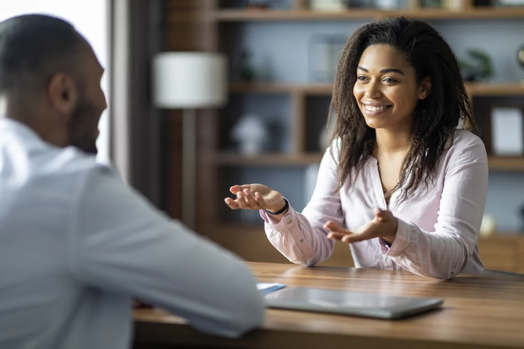 Dedicated permanent recruitment agency delivering expert recruitment services and recruitment outsourcing: Enthusiastic female consultant with curly hair in pink blouse, gesturing animatedly while engaging male candidate in white shirt at wooden desk with laptop and notebook, in cozy office with bookshelves, lamp, and plants, fostering successful talent hiring partnerships.