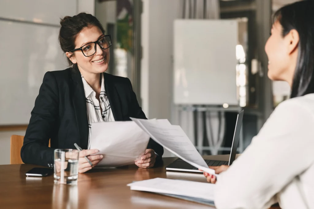 Local recruitment agency specialising in recruitment consulting: Smiling female consultant in glasses and black blazer reviewing documents with enthusiastic female candidate in white blouse at wooden desk with laptop, water glass, phone, and notebooks in modern office, driving personalised local talent acquisition and hiring success.