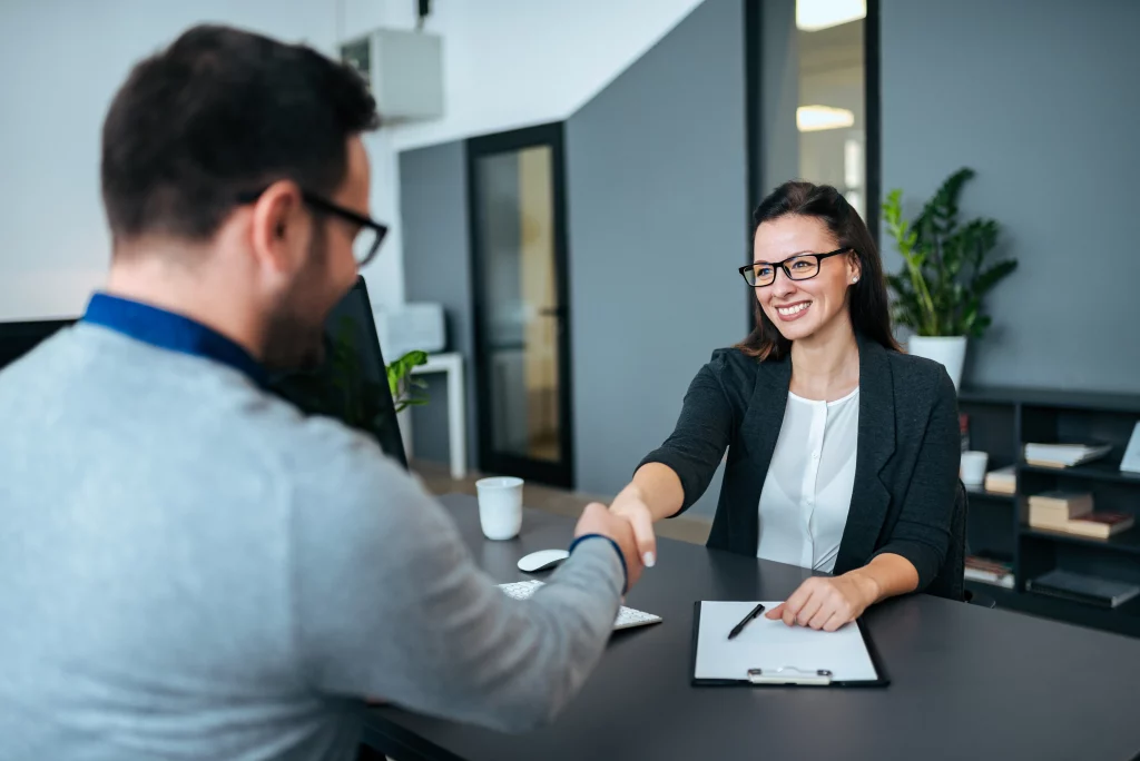 Expert recruitment agency delivering recruitment consulting and perm recruitment agencies expertise: Smiling female consultant in glasses and gray blazer warmly shaking hands with male job candidate in blue-collared gray sweater at modern office desk featuring clipboard, pen, mouse, coffee mug, and potted plants, building strong permanent talent acquisition connections.