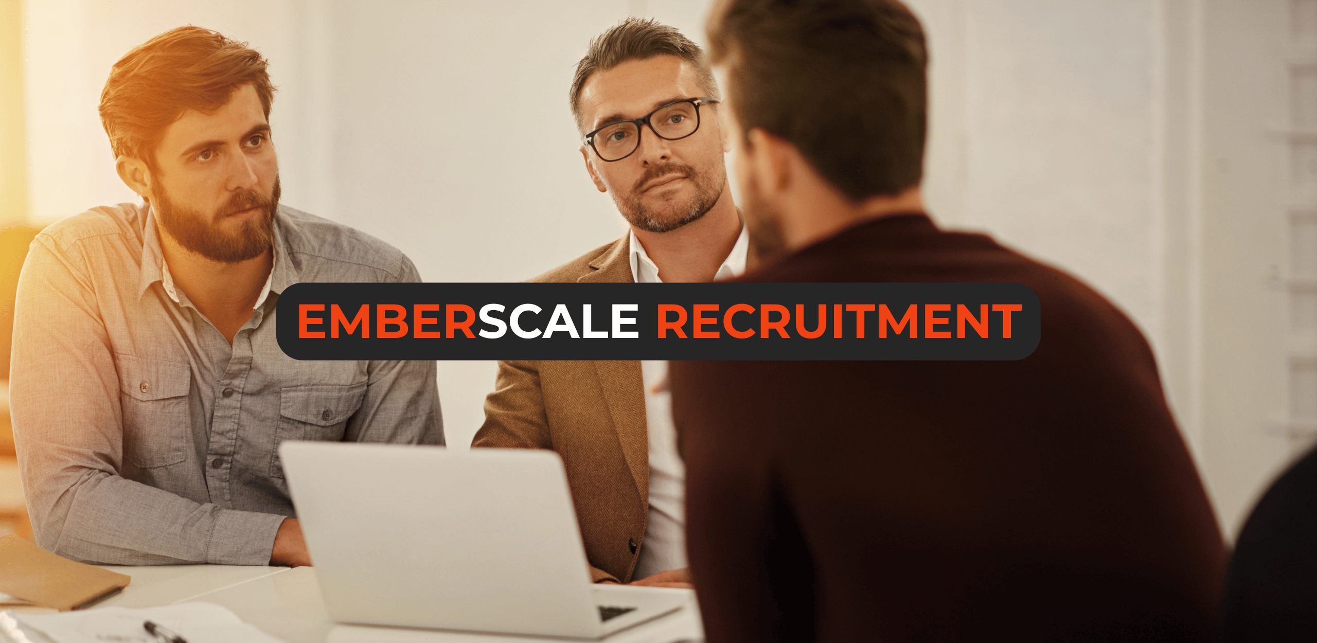 Two male hiring managers at a desk with a laptop interviewing a male candidate who was sourced by a recruitment agency