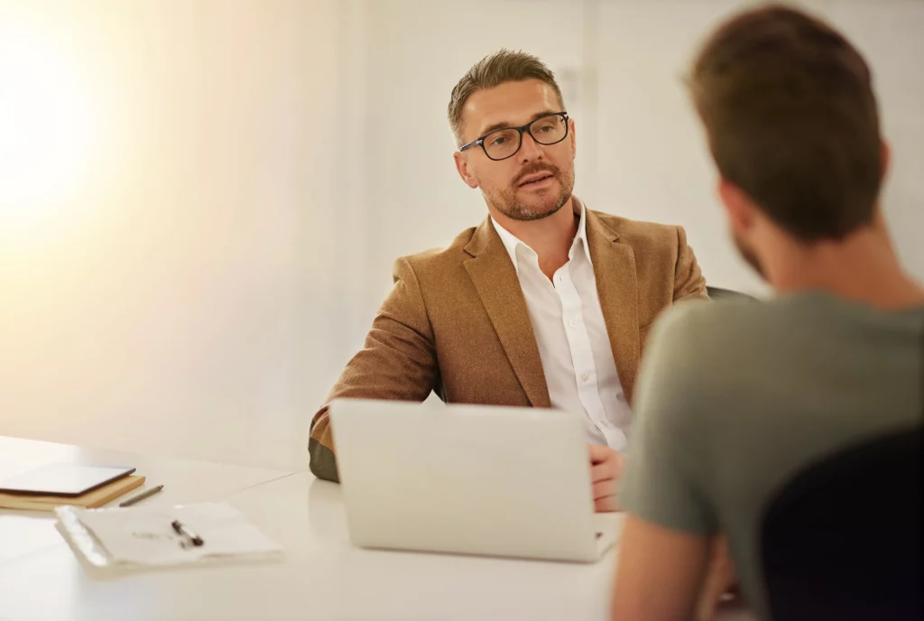 Professional recruitment agency consultant wearing glasses and a brown suit jacket, engaged in a one-on-one interview with a job candidate at a modern office desk featuring a laptop, notebooks, and documents, warm lighting creating an inviting atmosphere for talent hiring discussions.