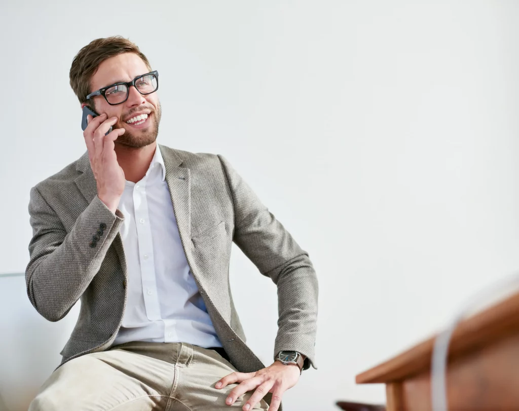 Confident RPO (Recruitment Process Outsourcing) expert delivering tailored recruitment for small businesses: Professional consultant in glasses, gray blazer, white shirt, and khaki pants, smiling while on a phone call in a modern office, fostering efficient talent sourcing and hiring solutions.