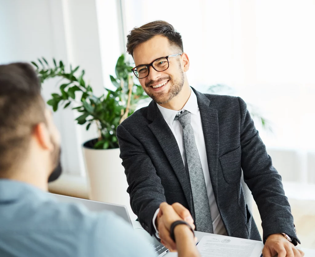 Expert perm recruitment agency consultant providing contingency recruitment agency services: Smiling male professional in glasses, gray suit, and tie, firmly shaking hands with job candidate in blue shirt at modern office desk with laptop, documents, and potted plant, embodying successful permanent and contingent talent acquisition partnerships.