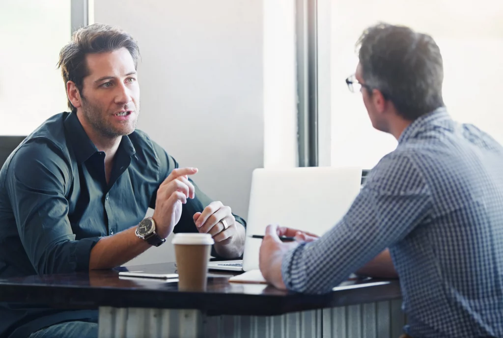 two men a manager in a formal meeting at a table with an employee implementing his training on how to dismiss an employee for poor performance