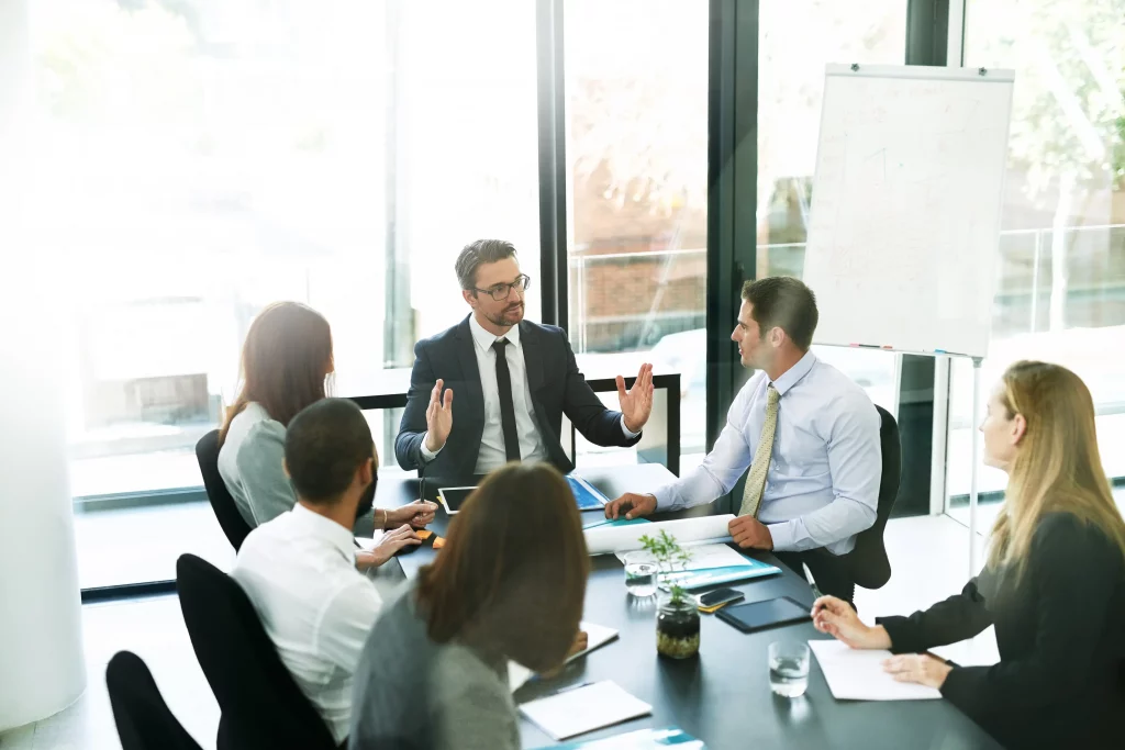 An SEO consultant in a suit presenting data analytics and upward-trending growth charts on a whiteboard to an attentive group of business professionals during a strategy meeting in a modern, brightly lit conference room.