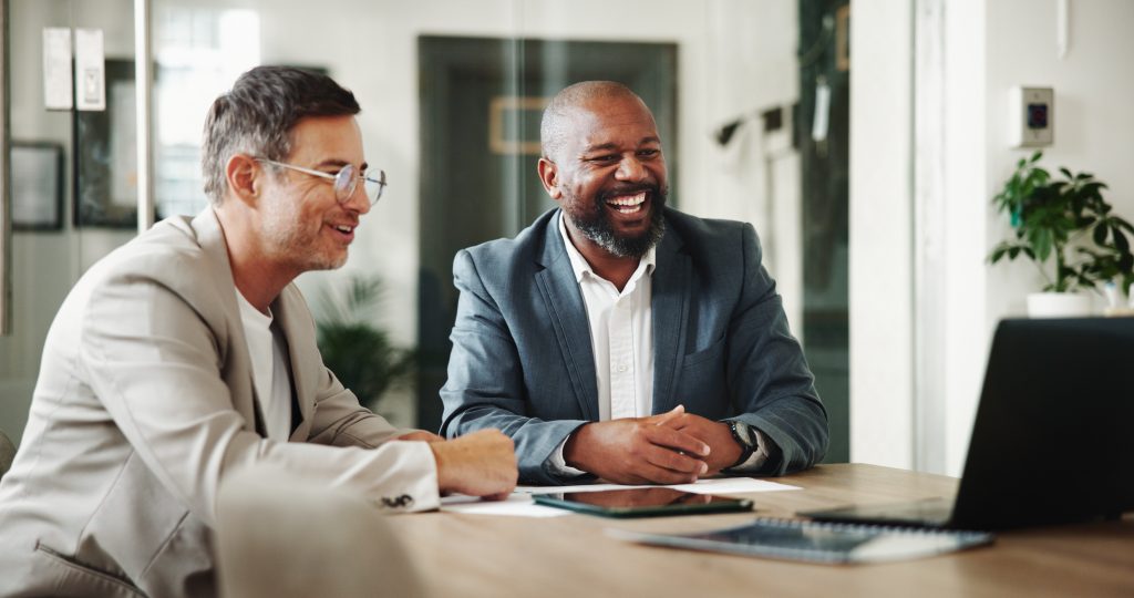 Two smiling business professionals in suits happily discussing affordable web design for small business during a consultation meeting with a laptop and documents on the table.