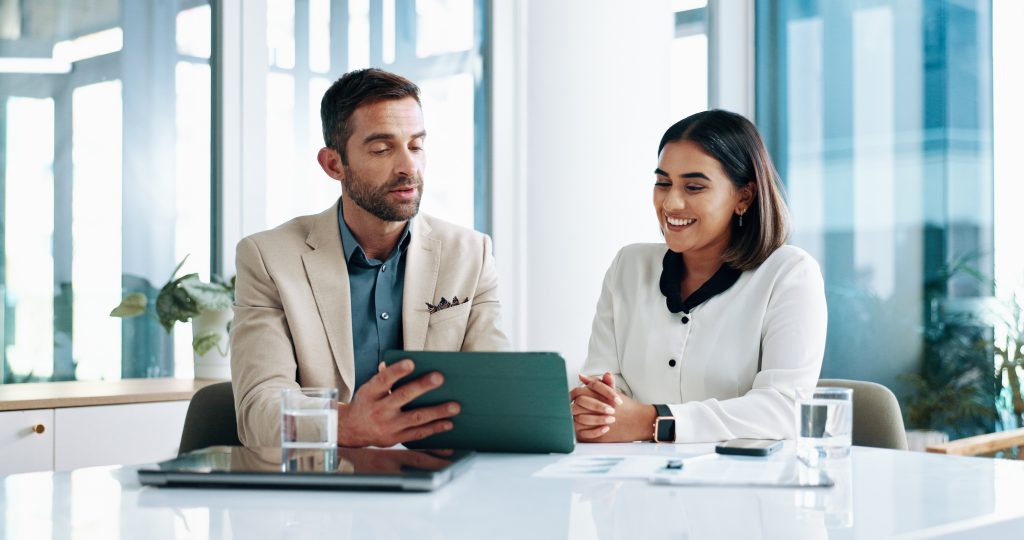 Two professional web designers smiling as they collaborate on a new website build, reviewing designs on a tablet during a meeting in a bright, modern office.